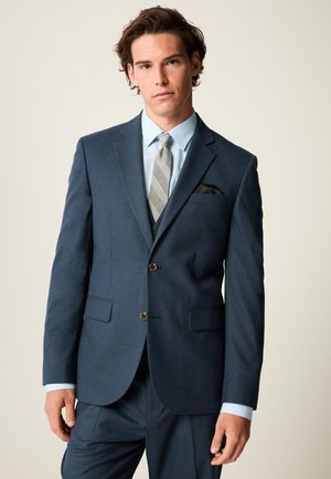 Mladý muž v námornícky modrom obleku so svetlomodrou košeľou a šedou vzorovanou kravatou na jednofarebnom svetlom pozadí.