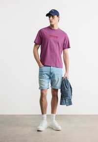 Νέος άνδρας που φοράει μοβ μπλούζα Calvin Klein, ανοιχτόχρωμο τζιν σορτς, λευκά αθλητικά παπούτσια και σκούφο ναυτικού, κρατώντας ένα μπλε πουκάμισο με καρό σχέδιο σε στούντιο.