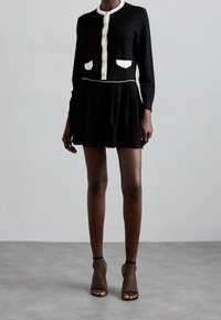 Kvinna står klädd i en svart klänning med vit kant, knappställd fram, och kort kjol, kombinerat med svarta högklackade skor med ankelremmar på en grå golv.