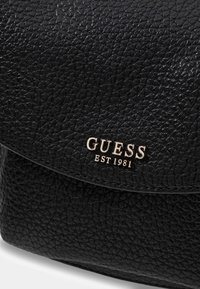 Zwarte leren tas met textuur en gouden "GUESS EST 1981" branding. Voorzien van een klep sluiting en een opvallend oppervlakpatroon. Compact ontwerp.
