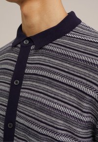Polo en maille avec col bleu marine, fermeture à boutons et motif rayé horizontal dans des tons bleu marine, gris et blanc.