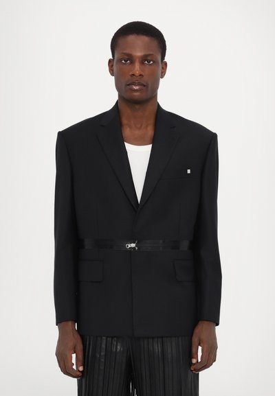 Black blazer with a structured design, flap pockets, a subtle logo detail, and a contrasting belt at the waist. Paired with pleated pants.