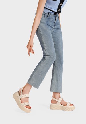 Persona che indossa jeans azzurri a gamba dritta e sandali beige con plateau e cinturini alla caviglia, camminando su uno sfondo bianco semplice.