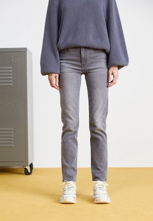 Person trägt einen violetten Strickpullover, graue Jeans und weiße Sneakers und steht drinnen auf einem gelben Boden neben einem grauen Spind.