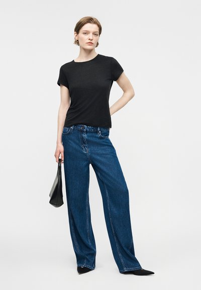Γυναίκα που φοράει μαύρο τοπ με κοντά μανίκια, μπλε τζιν παντελόνι με φαρδιά πόδια, μαύρα μυτερά παπούτσια και κρατάει μια μαύρη τσάντα, σε απλό ανοιχτό φόντο.