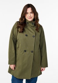 Cappotto doppiopetto verde oliva con bottoni neri, colletto alto, tasche laterali e taglio dritto. Tessuto liscio con una lucentezza moderata.
