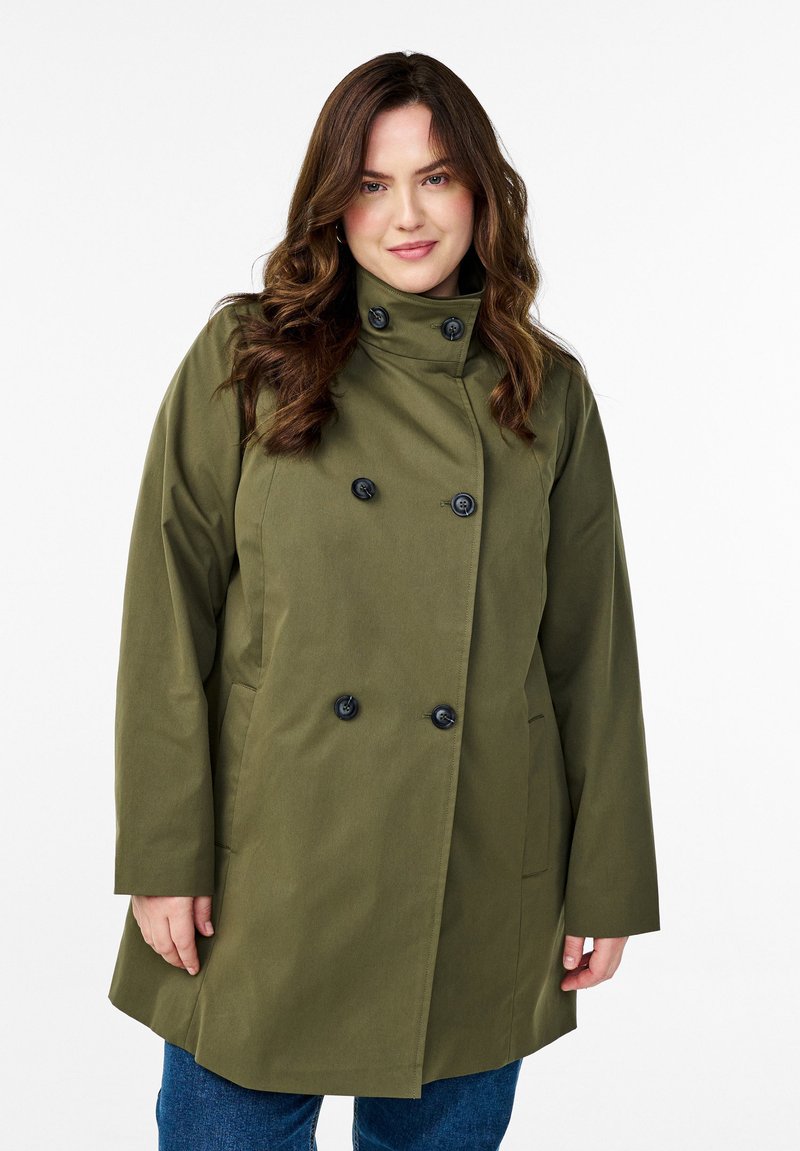 Cappotto doppiopetto verde oliva con bottoni neri, colletto alto, tasche laterali e taglio dritto. Tessuto liscio con una lucentezza moderata.
