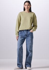 Olivgrön sweatshirt med ribbade ärmar, kombinerad med ljusblå, löst sittande jeans och vita sneakers, framför en enkel bakgrund.