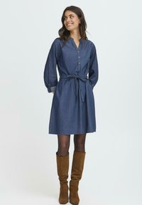 Kvinna som ler, klädd i en långärmad denimklänning med midjeband, svarta strumpbyxor och bruna knähöga suéde stövlar, stående framför en enfärgad bakgrund.