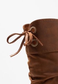 Botte en daim marron avec un revers roulé, dotée d'un lacet en haut et d'œillets en laiton pour l'ajustement. Texture lisse, design décontracté.