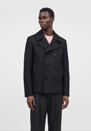 Homme portant un caban croisé noir sur une chemise rose et un pantalon noir ample, debout devant un fond clair uni.