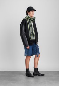 Melnā ādas un auduma pārstrādāta jaka, balts T-krekls, džinsu šorti ar sānu pogām, melni potīšu zābaki un zaļa akcentēta šalle.