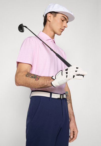 Vīrietis rozā polo kreklā un tumši zilos biksēs, tur golfu nūju pār plecu, valkā baltu cepuri un golfu cimdu, rādīdams roku tetovējumus.