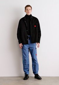 Camisa preta de botões sobre um suéter de gola alta preto, combinada com calças cargo azuis com bolsos laterais e detalhes em vermelho, usadas com sapatos pretos.
