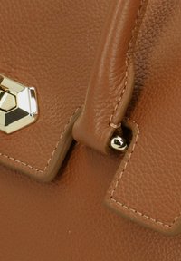 Angolo di una borsa a tracolla in pelle marrone texturizzata con un dettaglio hardware esagonale dorato e un accento di cucitura. Include un attacco a bottone metallico rotondo.