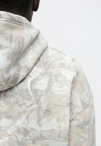 Felpa con cappuccio in beige chiaro con un motivo camouflage che presenta foglie e rami, tessuto morbido e design dalla vestibilità relax.