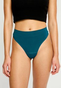 Femme portant un bas de bikini taille haute teal et un crop top noir, se tenant avec les bras détendus le long du corps devant un fond uni.