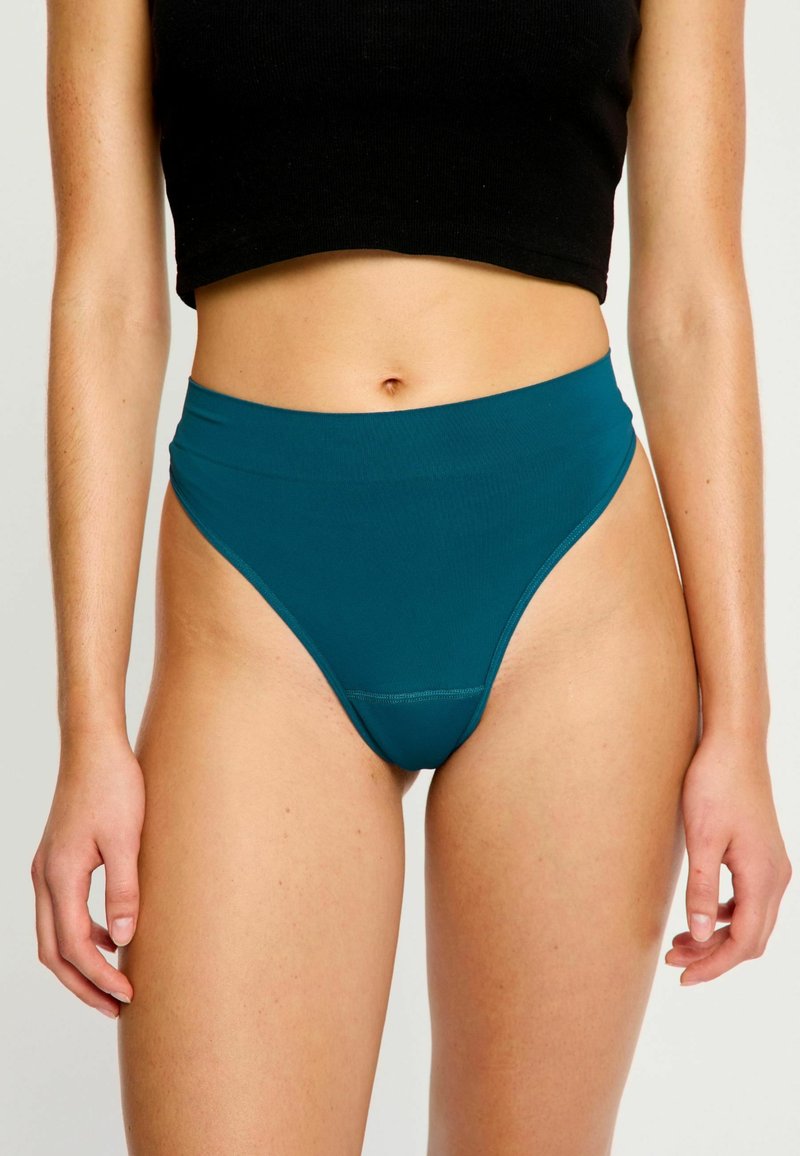 Femme portant un bas de bikini taille haute teal et un crop top noir, se tenant avec les bras détendus le long du corps devant un fond uni.