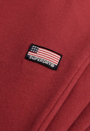 Czerwony materiał z naszytym prostokątnym łatą, na której znajduje się flaga USA oraz białym napisem "SUPERDRY®" na czarnym tle.