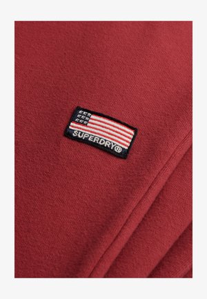 Czerwony materiał z naszytym prostokątnym łatą, na której znajduje się flaga USA oraz białym napisem "SUPERDRY®" na czarnym tle.