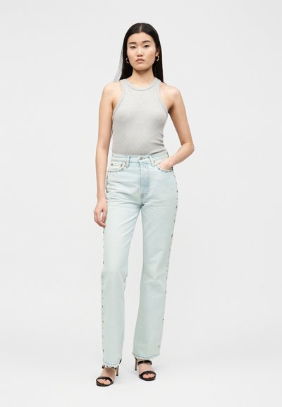 Femme aux longs cheveux noirs porte un haut gris sans manches, un jean taille haute bleu clair, des sandales noires à talons et des boucles d’oreilles créoles dorées, debout avec une main dans la poche.