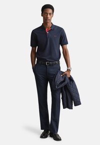 Tmavomodrá polo košeľa s krátkym rukávom a červeným golierom, čierny opasok, priliehavé tmavomodré nohavice, v ruke prešívaná tmavomodrá bunda. Čierne topánky dotvárajú vzhľad.