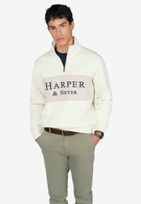 Sudadera blanca con cuello de media cremallera, que presenta una franja beige y letras en azul marino que dicen "HARPER & Neyer". Combinada con pantalones verde claro y un cinturón marrón.