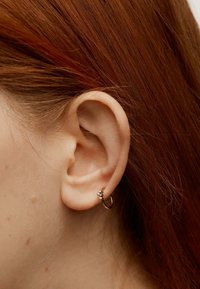 Pendiente de aro dorado con un pequeño grupo de piedras transparentes, llevado en una oreja con cabello rojo visible. Textura suave y diseño circular.