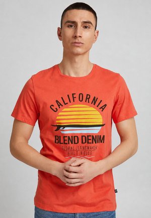 Muž v oranžovom tričku s nápisom „California Blend Denim“ a grafikou surfovej dosky, stojaci so zloženými rukami, neutrálny pozadie.