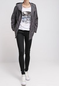 Grå hættetrøje med lynlås, hvid grafisk t-shirt, sorte skinny jeans og hvide sneakers; glat tekstur, afslappet design.