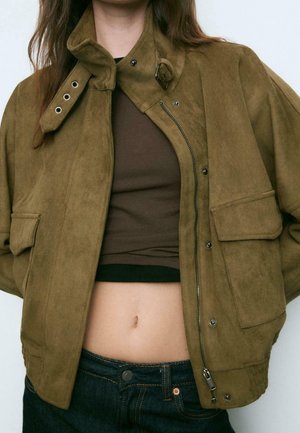 Personne portant une veste en daim vert olive dézippée avec de grandes poches par-dessus un haut marron court et un jean bleu foncé.