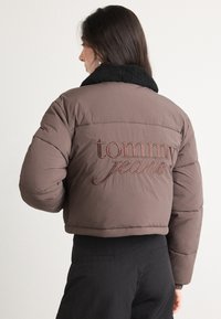 Brązowa kurtka puchowa z czarnym kołnierzem z owczej skóry. Posiada wyraźne haftowane logo "tommy jeans" w stylu pisma na plecach. Krótki fason.