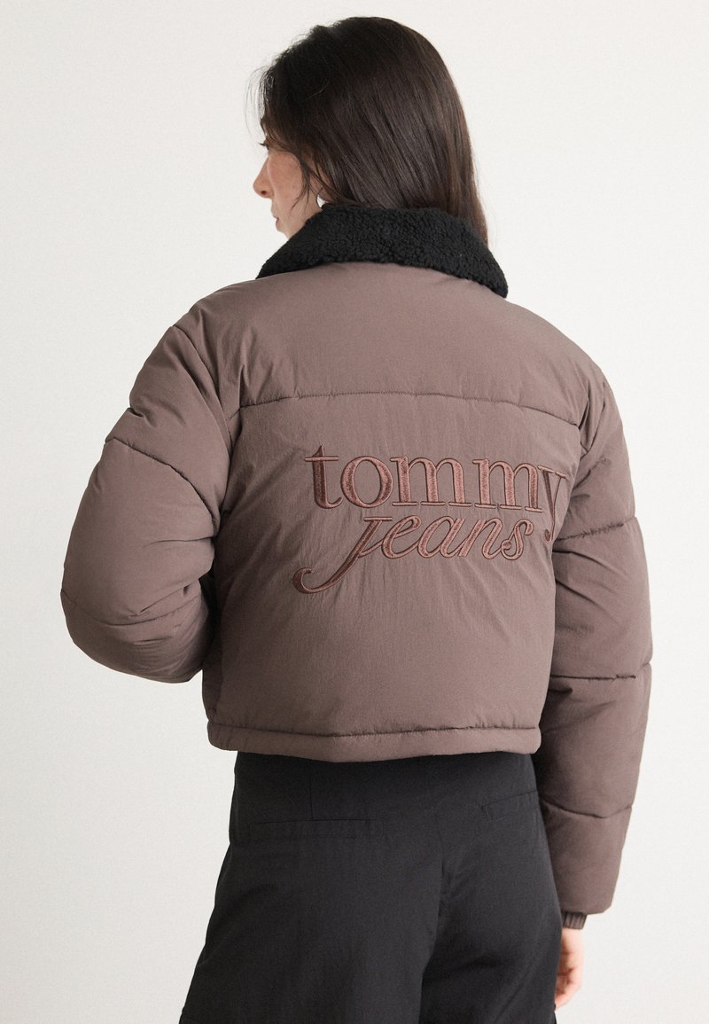 Brązowa kurtka puchowa z czarnym kołnierzem z owczej skóry. Posiada wyraźne haftowane logo "tommy jeans" w stylu pisma na plecach. Krótki fason.