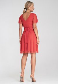 Rotes Kleid mit transparenten kurzen Ärmeln, offener Rückseite und gebundenem Taillenzug. Semiflared Rock, knielanger Saum, kombiniert mit Riemchenabsätzen.