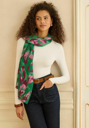 Sieviete ar cirtainiem matiem valkā baltu garā piedurkņa krekliņu, zaļu un rozā rakstainu šalli, tumšas džinsas ar brūnu jostu un koka aproces.