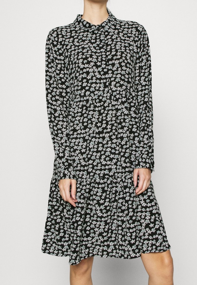 Robe noire avec un col, des manches longues et une jupe à volants. Présente un motif floral blanc sur toute la surface, fabriquée en tissu léger.
