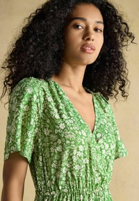 Grünes Blumenwickelkleid mit V-Ausschnitt, kurzen Ärmeln und elastischem Taillenbund. Das Kleid hat weiße Blumenmuster auf einem strukturierten Stoff.