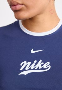Námořnické modré přiléhavé tričko s bílým žebrovaným límcem a centrálním bílým logem Nike v kurzívě. Na kůži je vidět malé srdíčkové tetování.