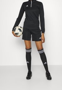 Personne en tenue de football Adidas noire tenant un ballon de football noir et blanc, portant des chaussettes hautes et des crampons, sur un fond uni.