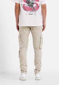 Beige cargobroek met meerdere zijzakken, taps toelopende benen en subtiele stiksels, gecombineerd met witte sneakers en een graphic t-shirt.