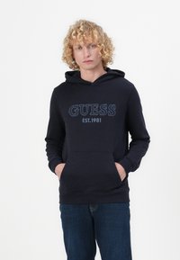 Felpa blu navy con tasca frontale, con la scritta "GUESS" e "EST. 1981" stampate in azzurro. Tessuto morbido e polsi a costine.