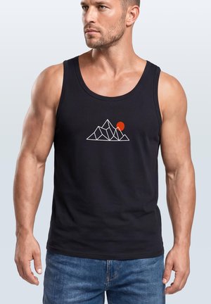 Man wearing black tank top with geometric mountain design and orange sun, standing against a plain light background.
