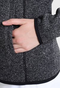 Hand tucked into side pocket of textured black and white herringbone jacket with black trim on sleeve and hem.