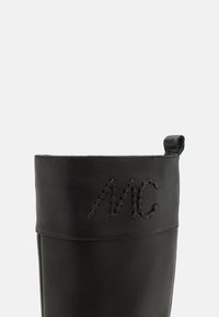 Botas de cuero negro con una textura suave y una forma redondeada. Presentan iniciales decorativas cosidas "MC" en el costado y una lengüeta.
