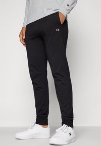 Sorte bomullssweatpants med en innsvingt design, elastiske mansjetter, sidelommer og liten brodert logo på venstre lår, kombinert med hvite sneakers.
