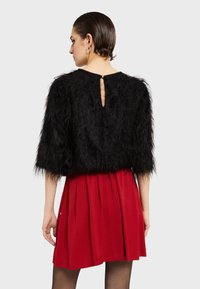 Čierny faux kožušinový cropped top s krátkymi rukávmi a detailom kľúčovej dierky na chrbte, skombinovaný so skladanou červenou sukňou. Hladká textura kontrastuje s nadýchaným materiálom.