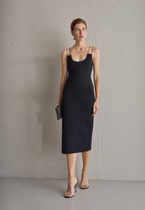 Γυναίκα σε εφαρμοστό μαύρο midi φόρεμα με λεπτές τιράντες, κρατώντας μια μικρή τσάντα, φορώντας ψηλές γόβες με λουράκια, στέκεται μπροστά σε μια υφασμένη γκρίζα τοιχοποιία.