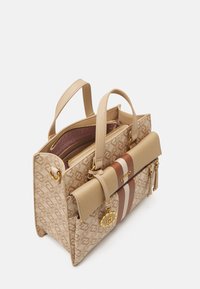Beżowa torebka o strukturalnym kształcie z monogramem, złotymi akcentami, odpinanym paskiem oraz kontrastującym brązowo-białym paskiem dekoracyjnym.