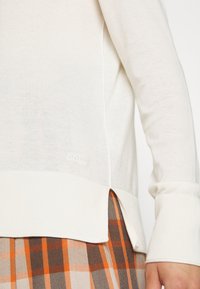 Krémový pletený svetr s vroubkovanými akcenty, bočními rozparky a jemným detailem loga. Kombinovaný s hnědým a oranžovým kostkovaným sukněmi.