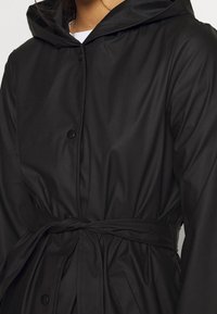 Svart vattentålig jacka med huva, knäppning med knappar, knytbälte i midjan och slät yta. Enkelt design, inga synliga mönster eller accentuationer.